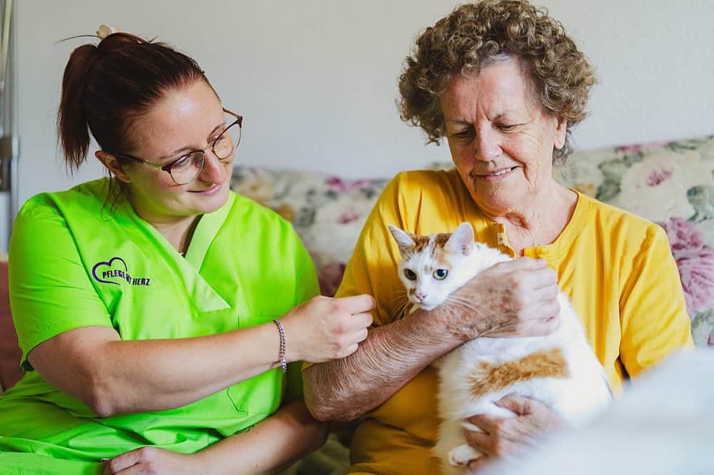 Pflegekraft in grünem Kasack krault die Katze, die eine ältere Dame auf ihrem Sofa im Arm hält