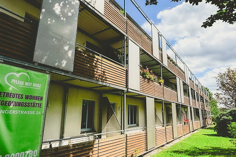 Wohnhaus Klingerstraße 26 mit Holzbalkonen voller Blumenkästen, davor das grüne Schild von Pflege mit Herz