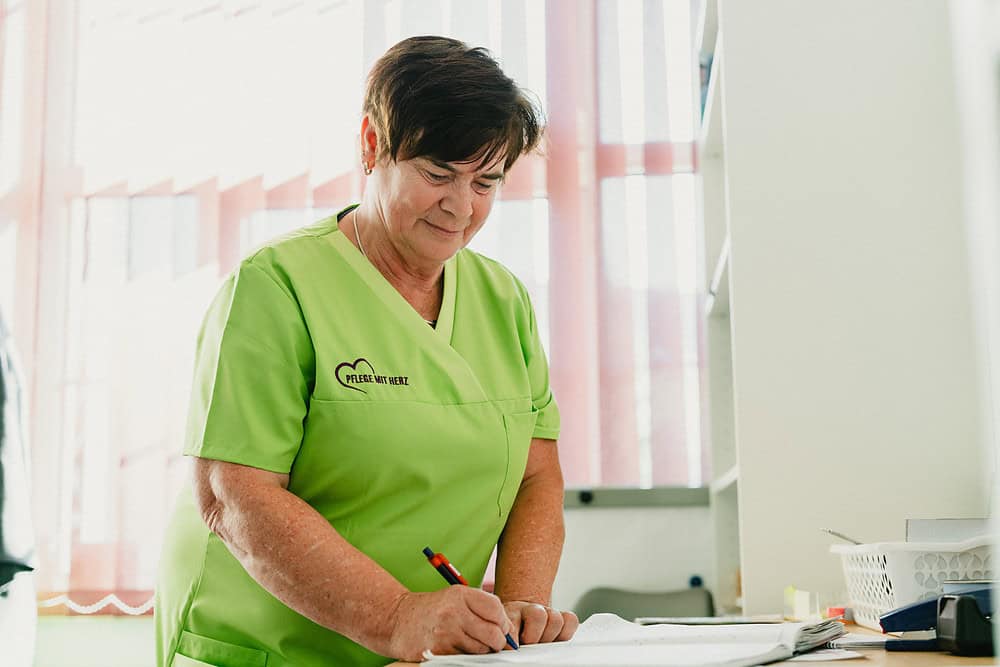 Pflegekraft im hellgrünen Kasack schreibt am hellen Fenster in die Pflegedokumentation