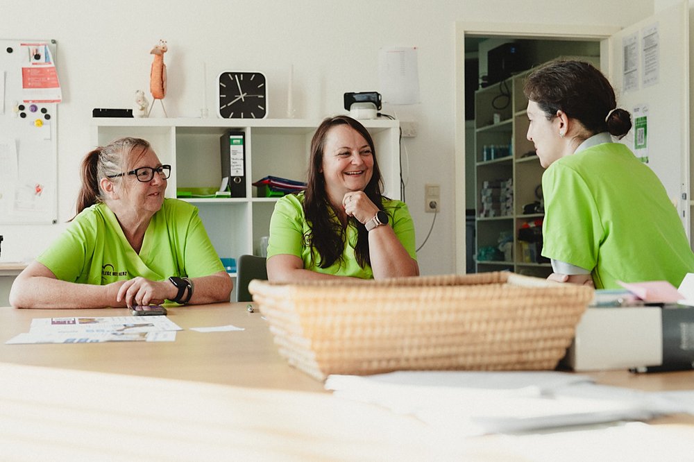 Drei Mitarbeiterinnen in grünen Poloshirts sitzen lachend an einem Tisch im hellen Büro und besprechen sich